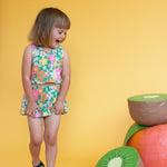 Young girl in a floral dress standing next to oversized fruit props on a yellow background