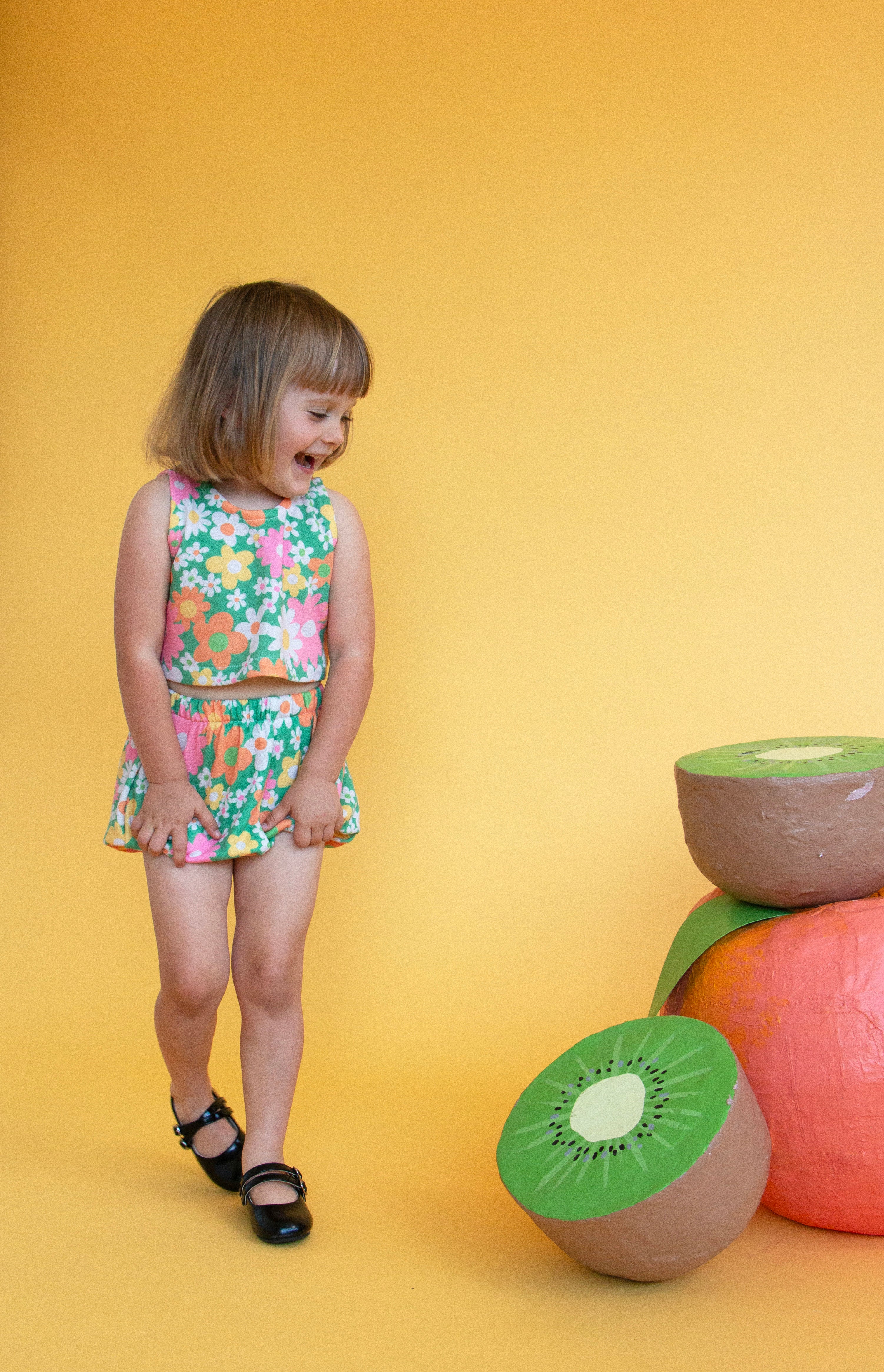 Young girl in a floral dress standing next to oversized fruit props on a yellow background