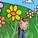 Child standing in front of a colorful mural of flowers and greenery wearing 80s ringer tee