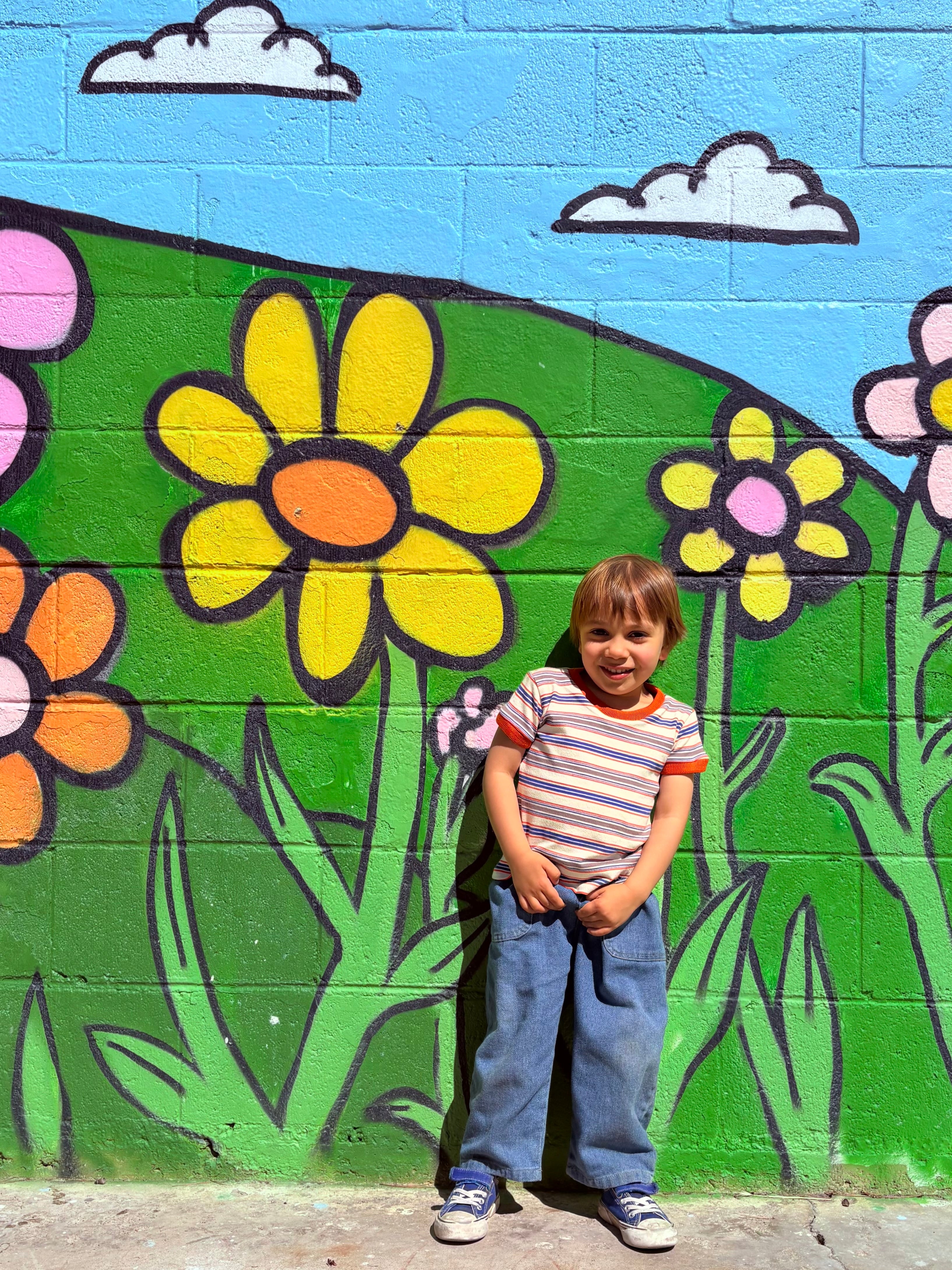 Child standing in front of a colorful mural of flowers and greenery wearing 80s ringer tee