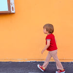 Child in a red shirt and checkered pants walking on a sidewalk with an orange wall in the background.