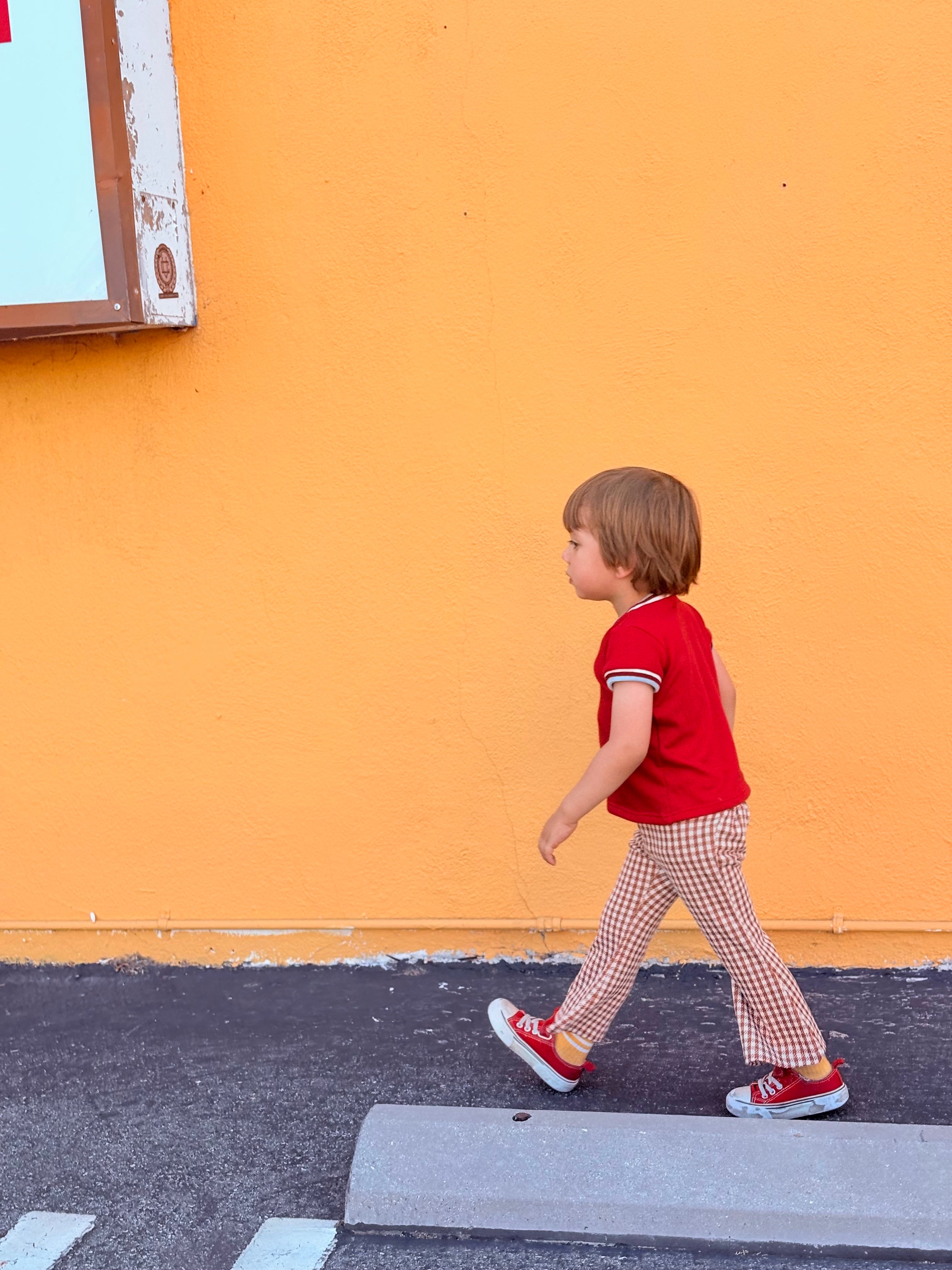 Child in a red shirt and checkered pants walking on a sidewalk with an orange wall in the background.