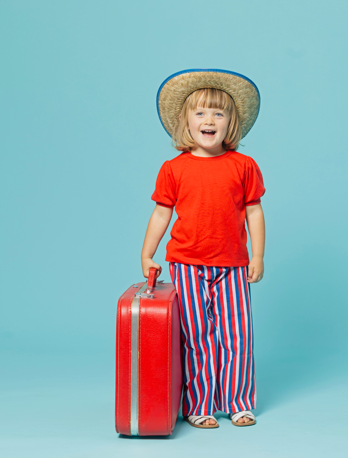 child wearing cool outfit with red shirt, vintage style wide leg striped pants, holding a red suitcase and a blue background