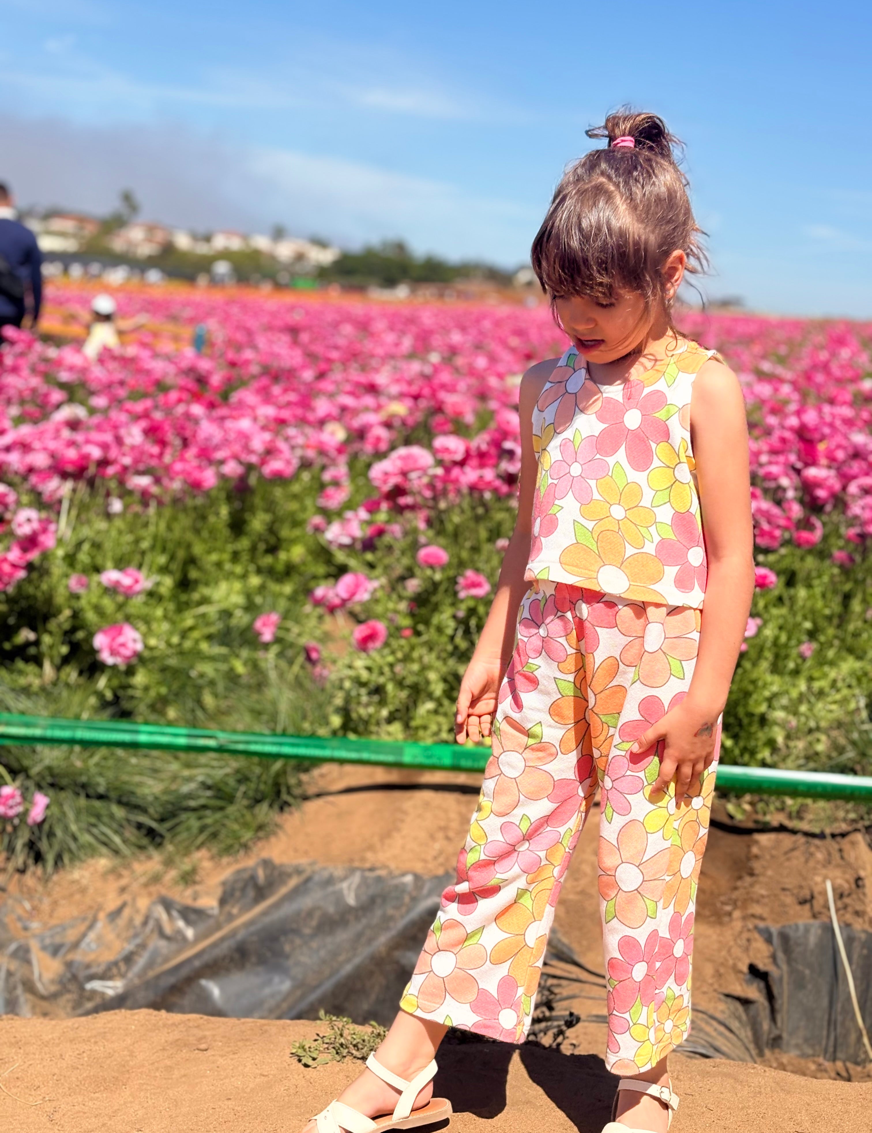 Child in a colorful flower power birthday outfit standing in a field of pink flowers with a clear blue sky.
