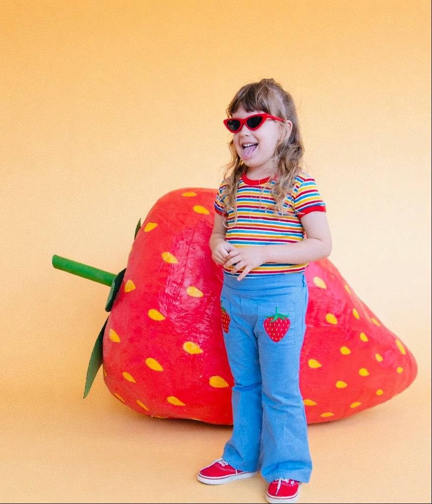 Girl wearing fun colorful vintage inspired striped shirt and strawberry pants in front of giant strawberry prop 