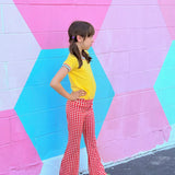 Child wearing a yellow shirt and red retro checkered bell bottom pants standing in front of a colorful geometric-patterned wall.