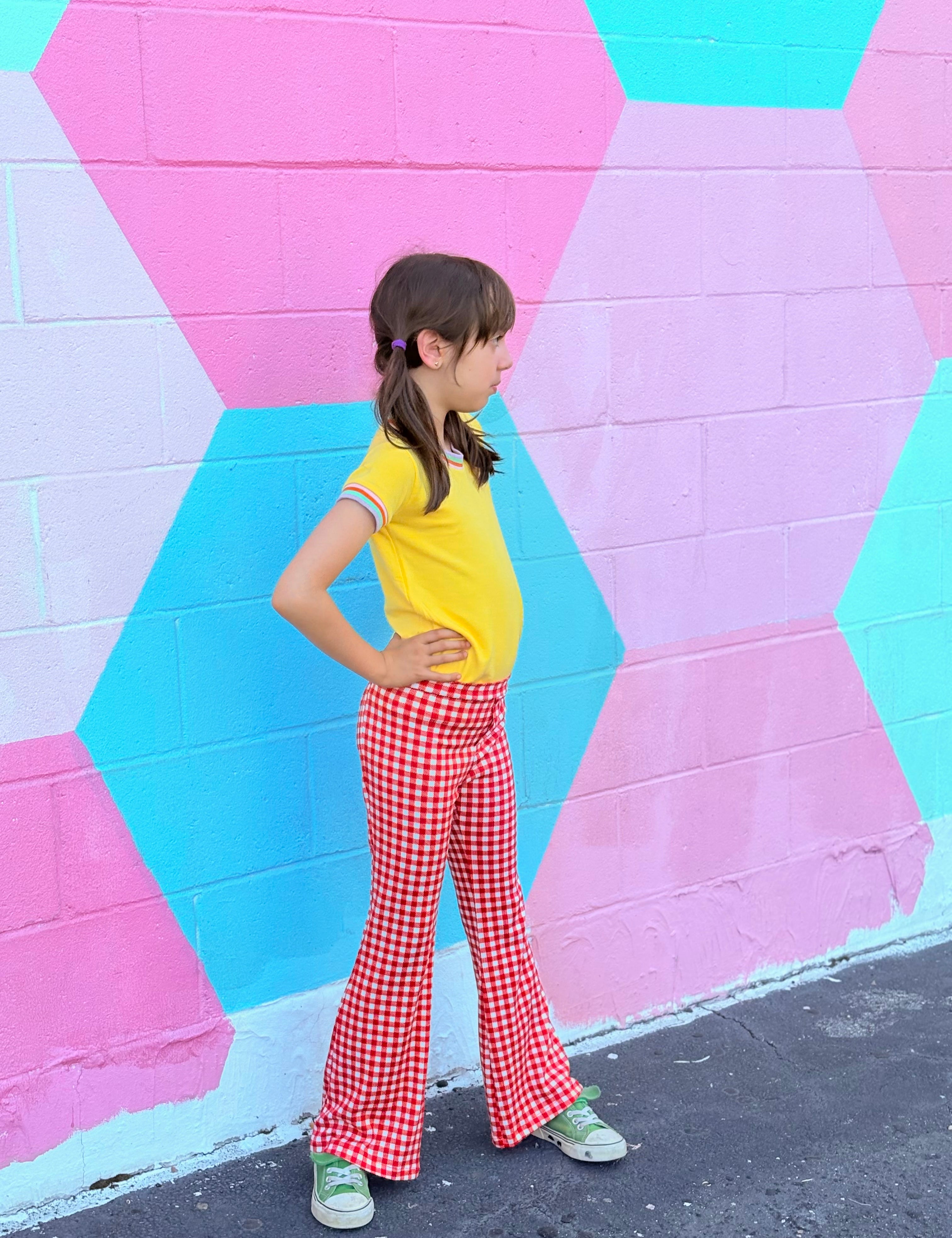 Child wearing a yellow shirt and red retro checkered bell bottom pants standing in front of a colorful geometric-patterned wall.