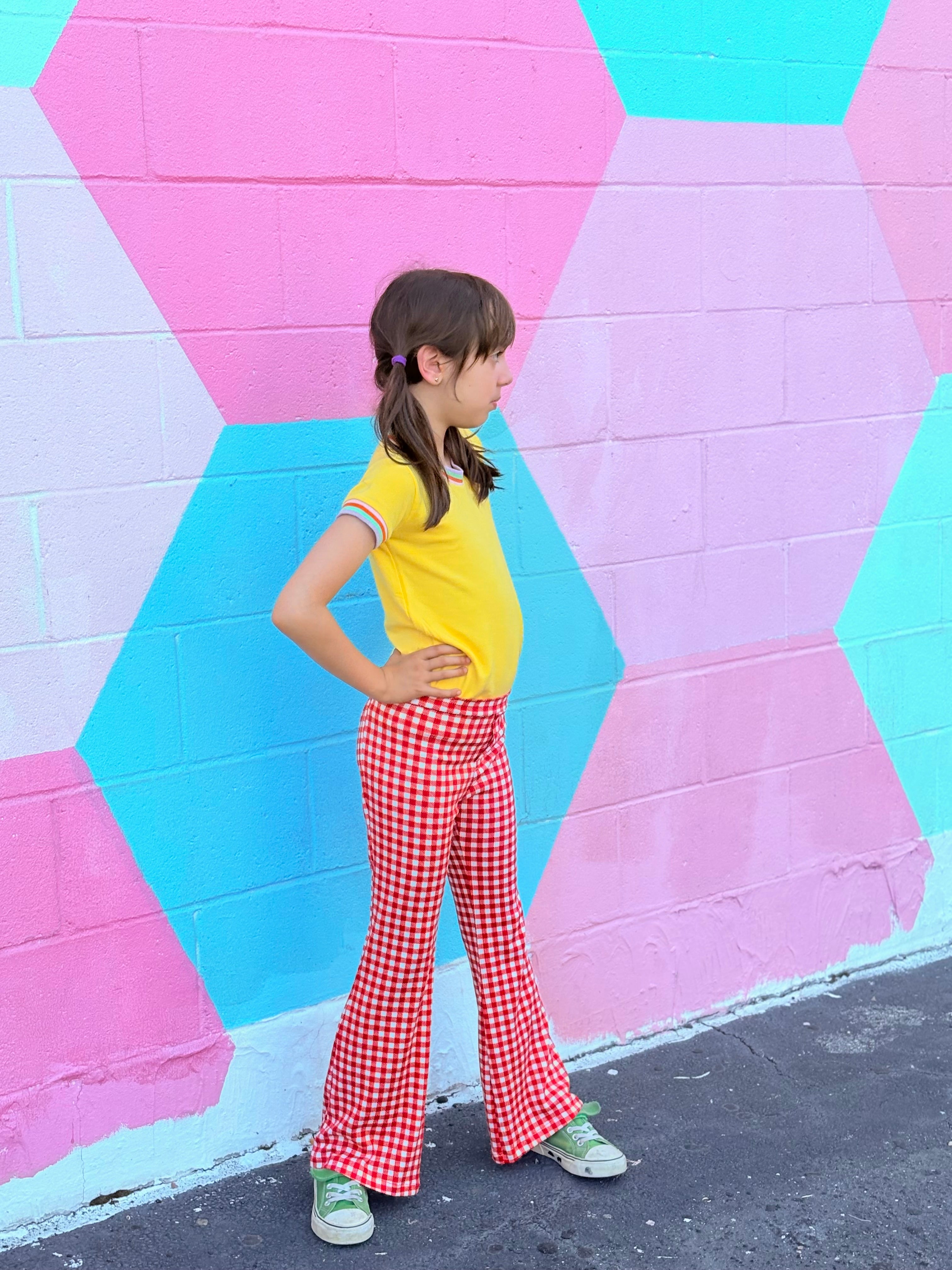 Child wearing a yellow shirt and red retro checkered bell bottom pants standing in front of a colorful geometric-patterned wall.