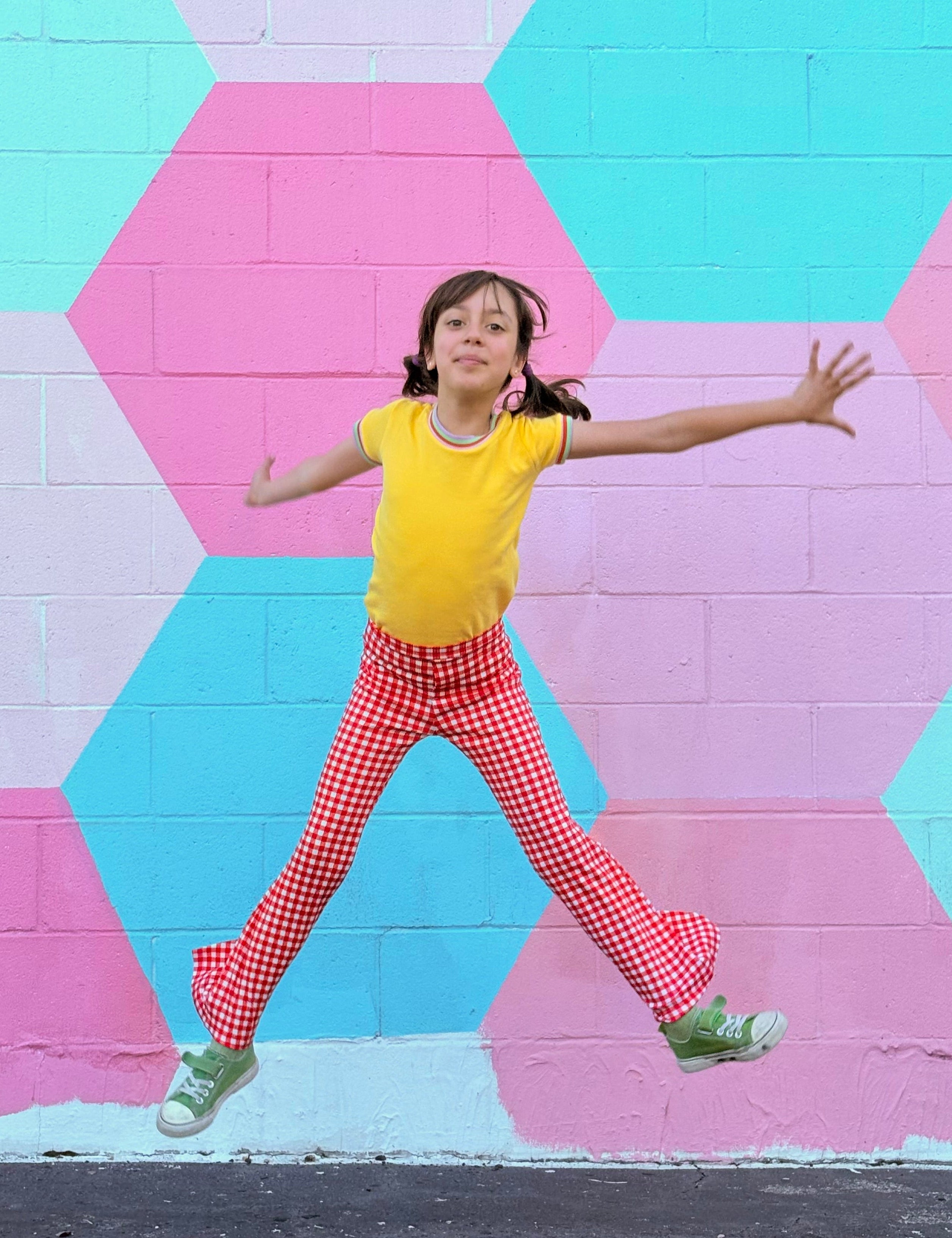Child in yellow shirt and red checkered bell bottom pants jumping in front of a colorful geometric-patterned wall.