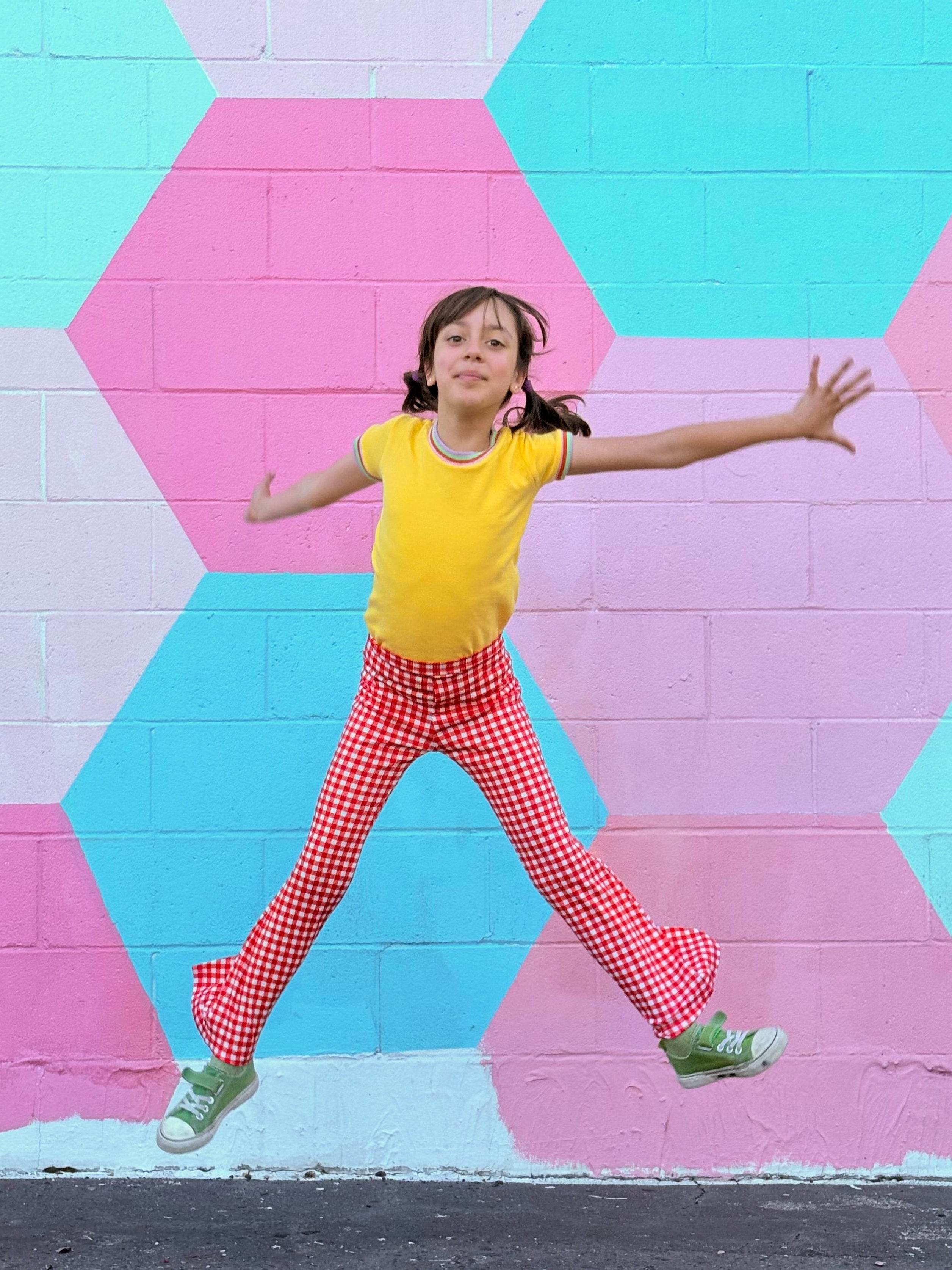 Child in yellow shirt and red checkered bell bottom pants jumping in front of a colorful geometric-patterned wall.