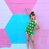 Young girl in a green romper with white floral patterns standing against a colorful geometric-patterned wall.