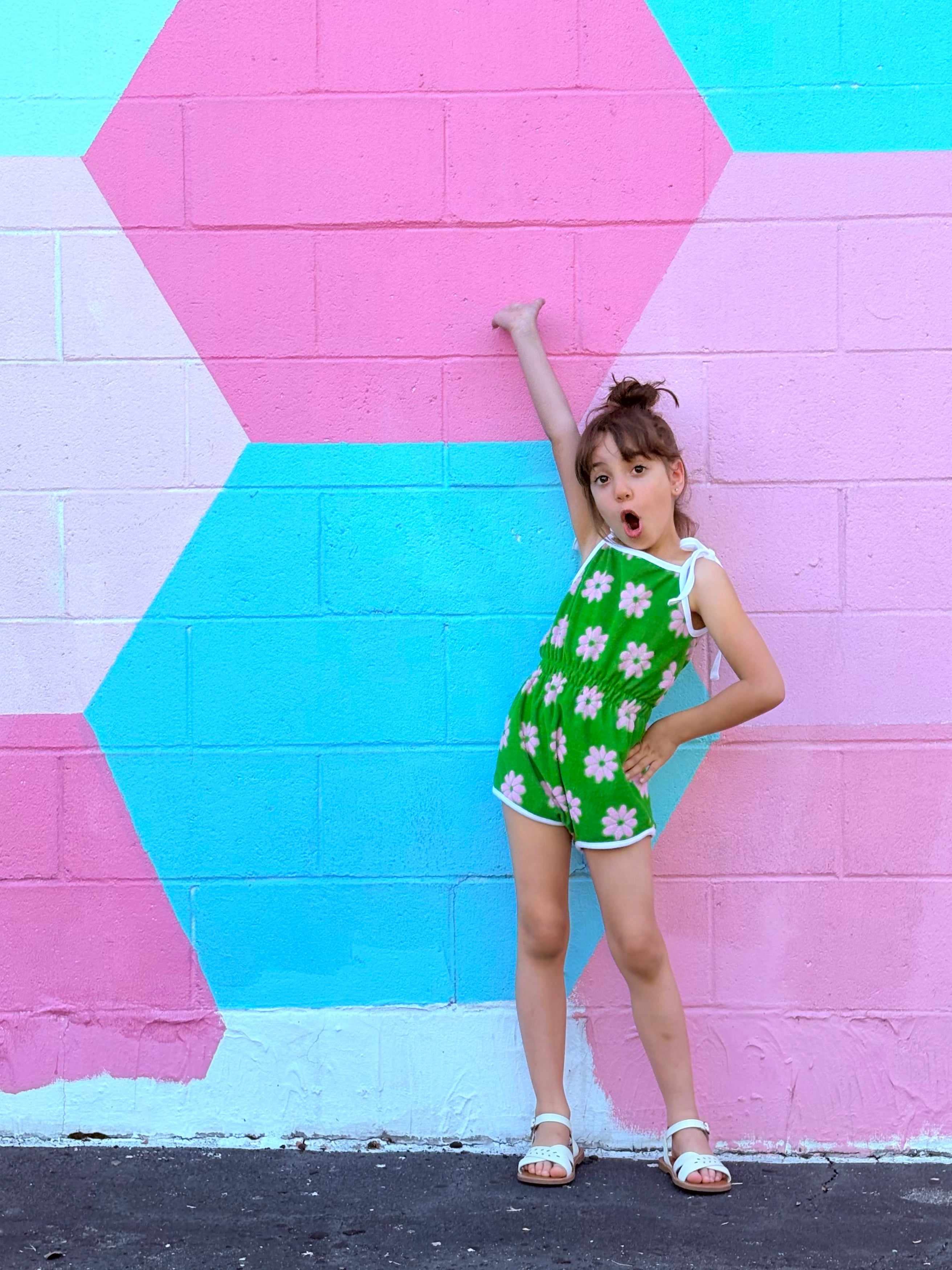 Young girl in a green romper with white floral patterns standing against a colorful geometric-patterned wall.