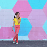 Girl wearing funky retro yellow top and red checkered pants standing in front of a colorful geometric-patterned wall.