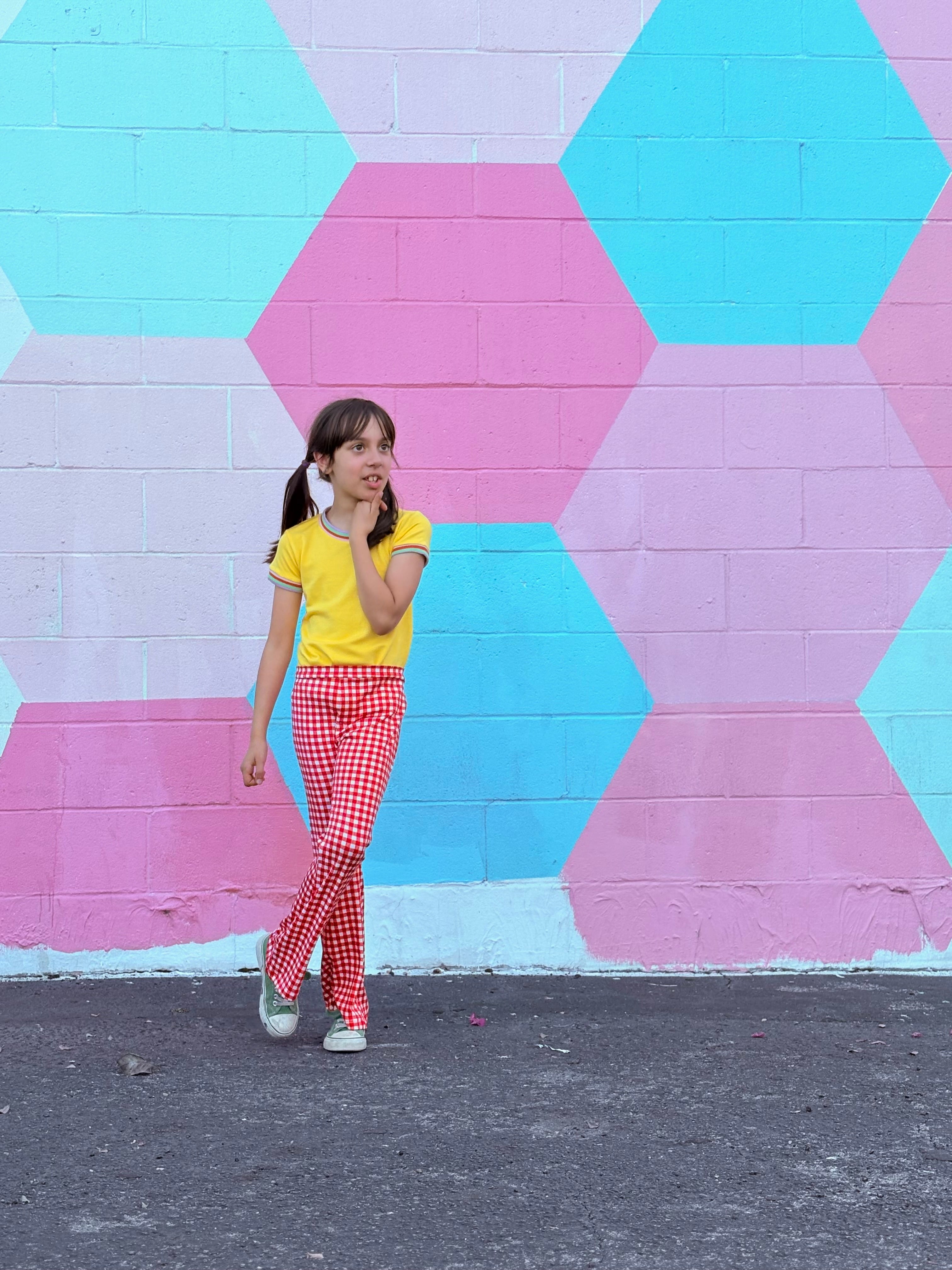 Girl wearing funky retro yellow top and red checkered pants standing in front of a colorful geometric-patterned wall.