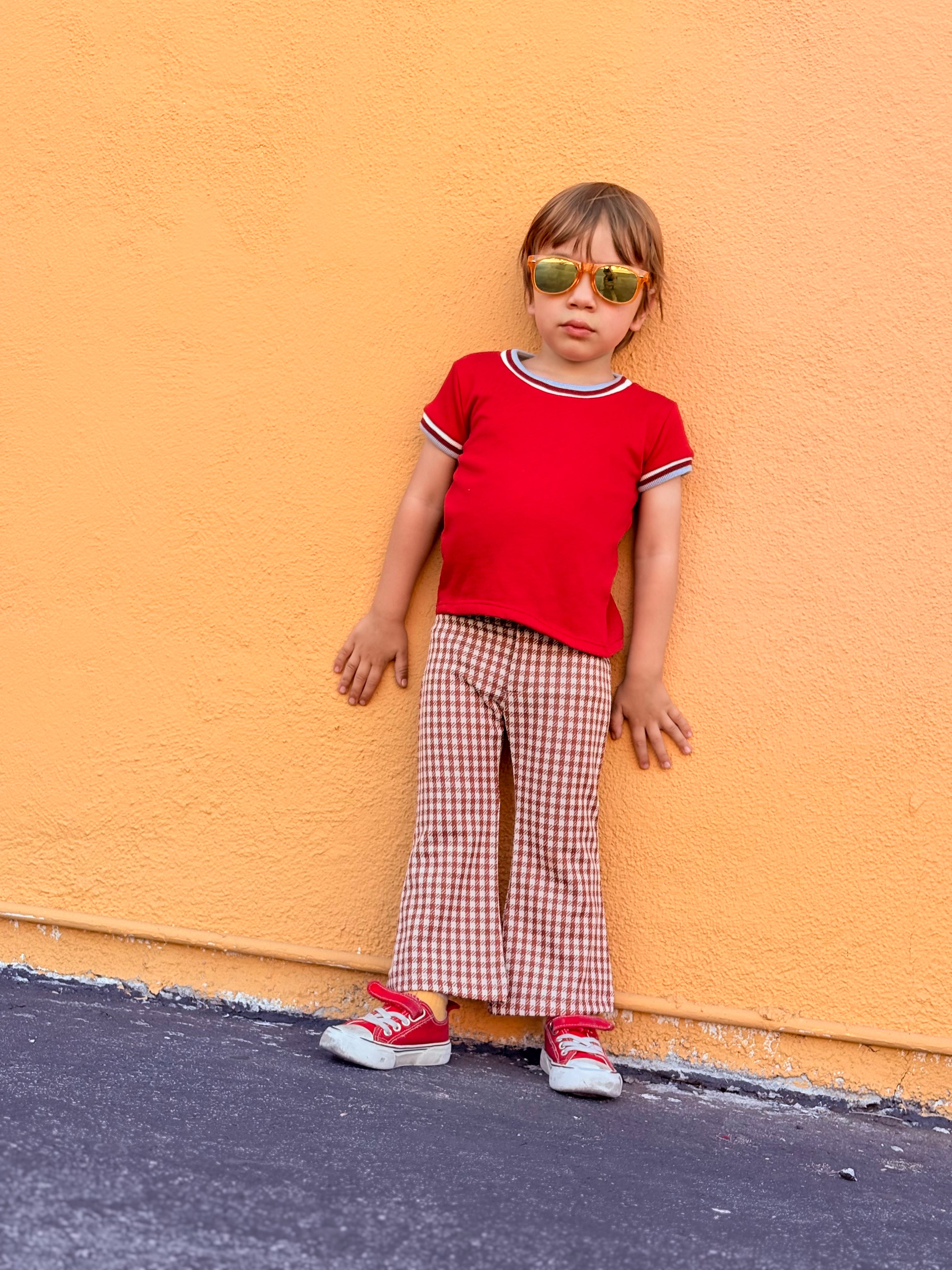 Child wearing a red shirt and checkered bell bottom pants against an orange wall